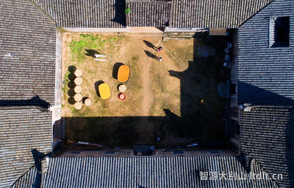 航拍下的老屋天井，秋日暖阳把屋檐影子拉长，院中竹匾摊着新晒的收成，两个人在阳光里忙碌